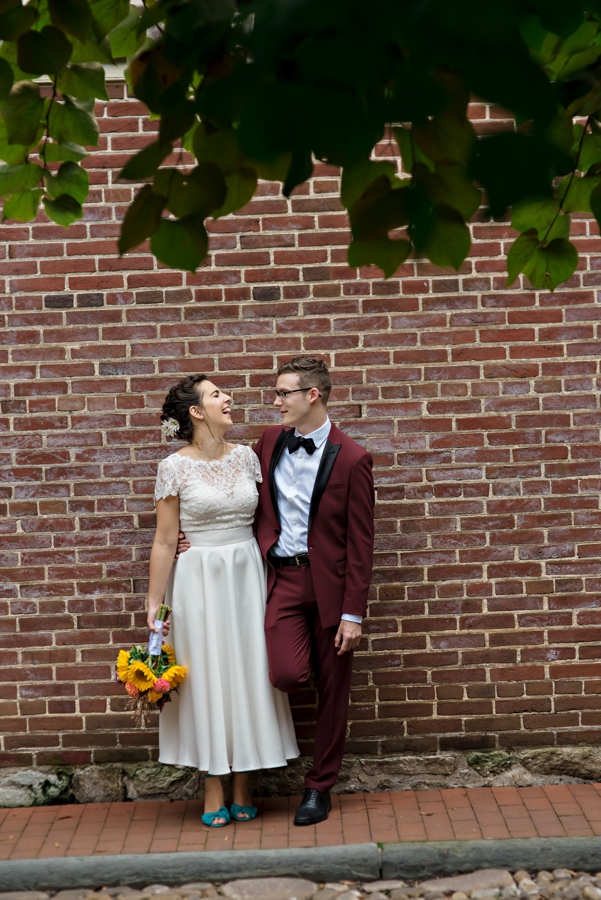 A bride and groom stand agains a brick wall under a green tree.